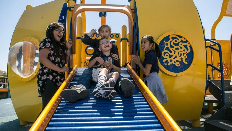 A child going down a slide at an inclusive playground guided by two other children and an adult.