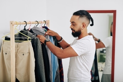 Handsome bearded man looking at clothes on hangers and choosing what to wear