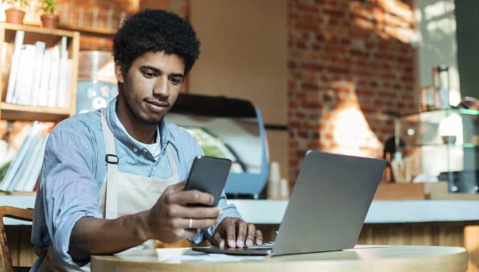 man with apron looks at cellphone and laptop