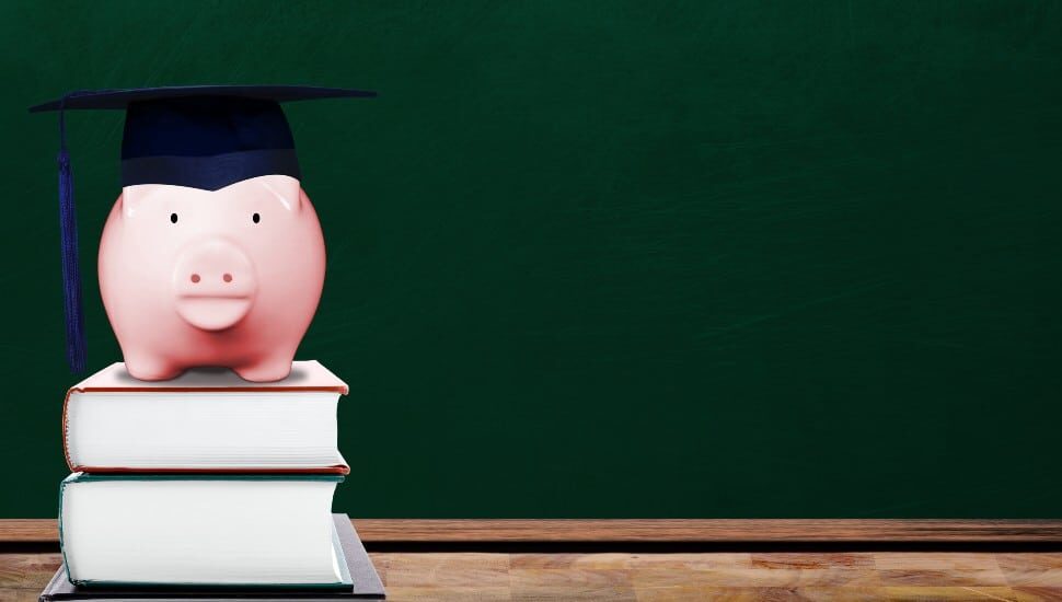 Saving for education with pink piggy bank wearing graduate hat on books in school classroom with copy space on chalkboard. Concept of education investment, tuition cost, college loan, etc.