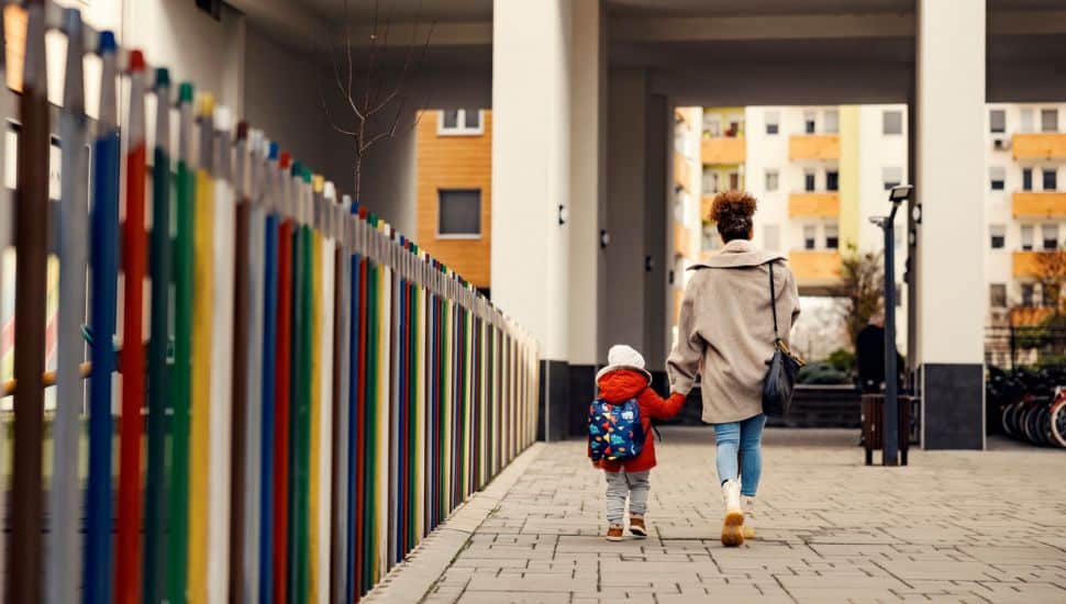 A young woman holding hands with little three year old boy with a schoolbag and taking him to kindergarten.