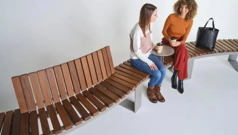 Two women with coffee talking while seated on a DuMor bench.
