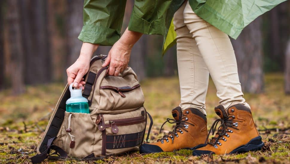 Woman with raincoat and hiking boots taking out water bottle from backpack.