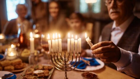 Close up of extended Jewish family celebrating Hanukkah at dining table. Focus is on mature man lighting candles in menorah.