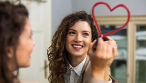 a young woman drawing a heart on a mirror with lipstick.