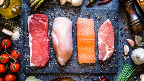 Top view of four different types of animal protein like a raw beef steak, a raw chicken breast, a raw salmon fillet and a raw pork steak on a stone tray. Stone tray is at the center of the image and is surrounded by condiments, spices and vegetables.