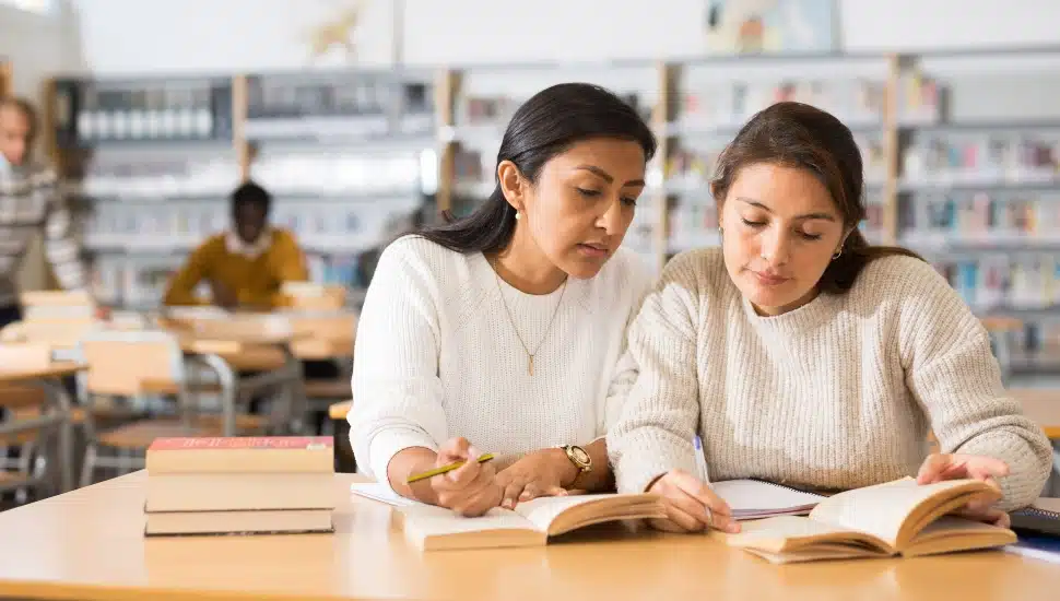 Two young adult women gaining new skills at public library, reading books and making notes.