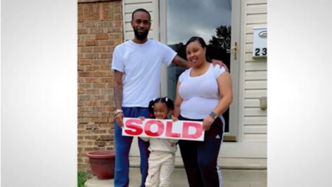 A couple and their young daughter in front of their new home holding a "Sold" sign.