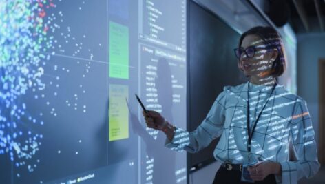 A young woman teacher gives a data science presentation with a projecting slide showing Artificial Intelligence Neural Network Architecture.