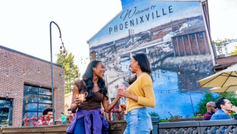 Two woman holding drinks talking in front of the Welcome to Phoenixville sign.