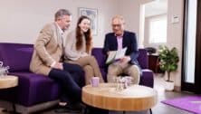 Customers sit on a couch inside a Propell Credit Union branch, engaged in a friendly discussion with a banker. The banker, smiling and attentive, explains financial options while holding documents. The setting is warm and welcoming, with modern furnishings and a community-focused atmosphere. The customers appear relaxed, asking questions and reviewing materials.