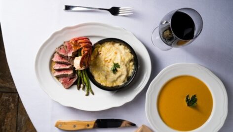 The image shows a sophisticated dinner setup at Taste & Sea in Chadds Ford featuring a surf-and-turf plate. The main dish includes slices of medium-rare steak served alongside a lobster tail and grilled asparagus. In the center of the plate is a small dish of creamy mashed potatoes, garnished with a sprig of parsley. Accompanying the meal is a bowl of creamy, orange-colored soup, also topped with parsley. To the right, a glass of red wine sits elegantly next to the plate, completing the upscale dining presentation. The table is set with a white tablecloth, a fork, and a knife with a wooden handle.