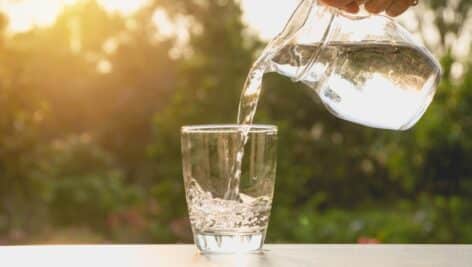 Person pouring water from pitcher to glass on nature background