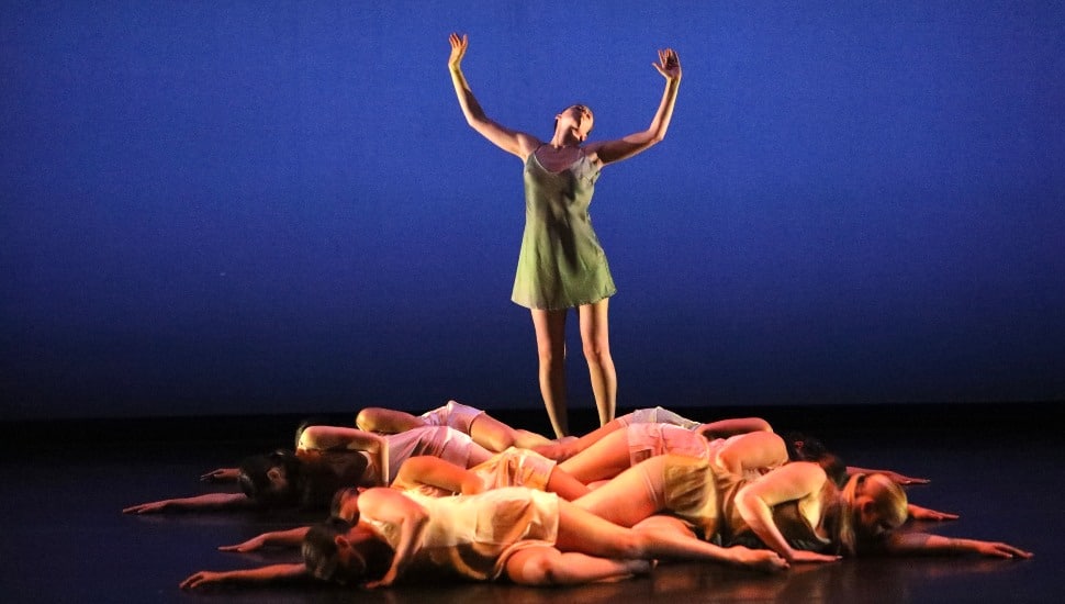 Dancers on stage at WCU. A dancer stands center stage in a powerful pose, wearing a green dress with arms extended upward, as if reaching toward the sky. She is illuminated against a deep blue background, which creates a dramatic and intense atmosphere. Surrounding her on the floor are several other dancers, lying in a circle around her, dressed in soft, neutral-colored costumes. They are positioned with their arms outstretched, creating a sense of unity and contrast with the standing figure, who appears to be the focal point of the scene. The overall effect is one of grace, strength, and harmony.