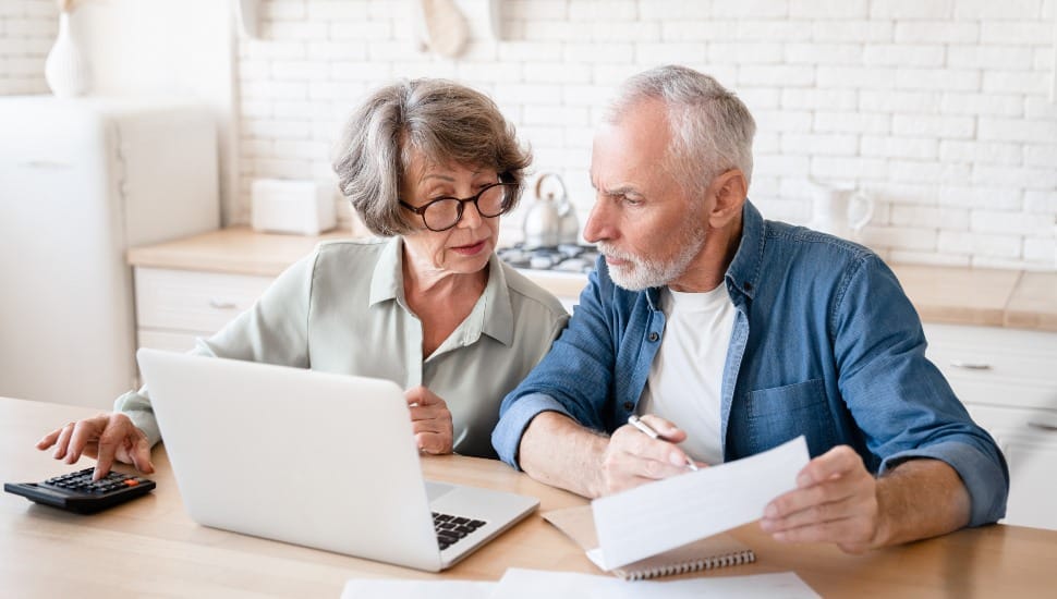 A senior couple sitting at a kitchen table or office desk with a laptop and paperwork, looking engaged and discussing Medicare options.