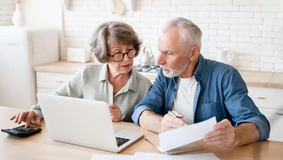 A senior couple sitting at a kitchen table or office desk with a laptop and paperwork, looking engaged and discussing Medicare options.