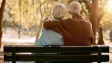 Elderly couple sitting on bench outside hugging each other.