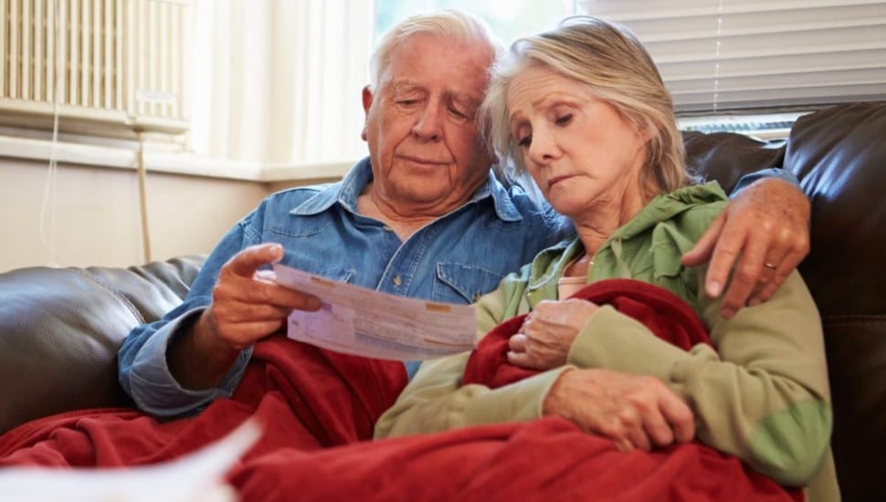 An elderly couple sitting together on a couch, looking concerned while reviewing a bill. They are wrapped in blankets, suggesting they are trying to stay warm indoors. The atmosphere conveys a sense of worry or financial strain, often associated with making tough decisions about heating or other household expenses.