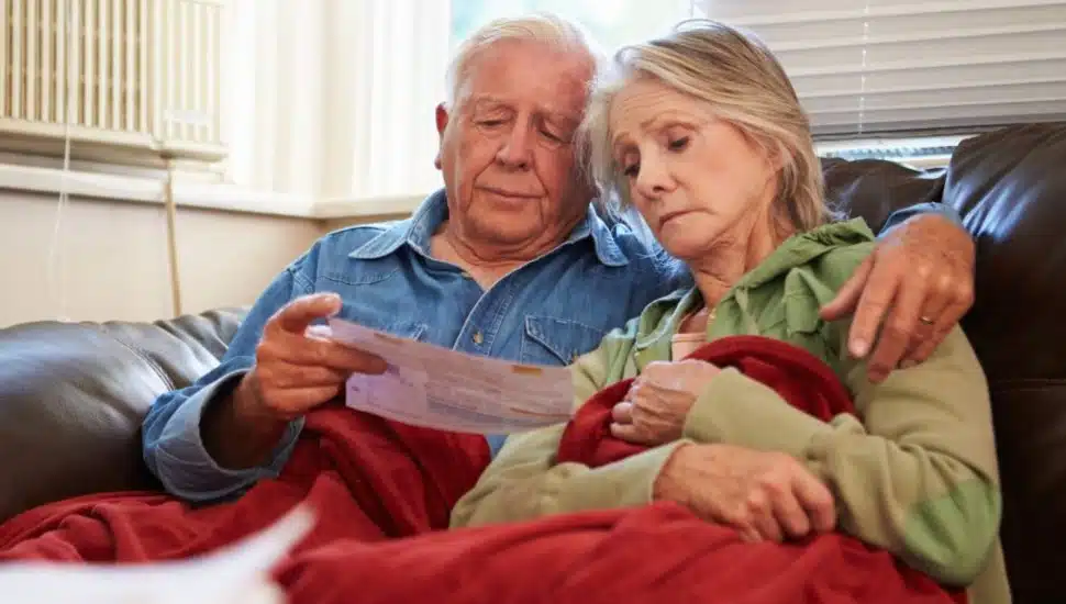 An elderly couple sitting together on a couch, looking concerned while reviewing a bill. They are wrapped in blankets, suggesting they are trying to stay warm indoors. The atmosphere conveys a sense of worry or financial strain, often associated with making tough decisions about heating or other household expenses.