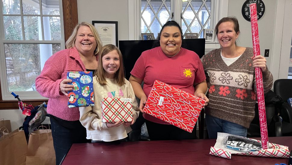 Two women and a girl hold wrapped gifts going to Chester County families. A third woman has a roll of wrapping paper about to wrap a donated gift.