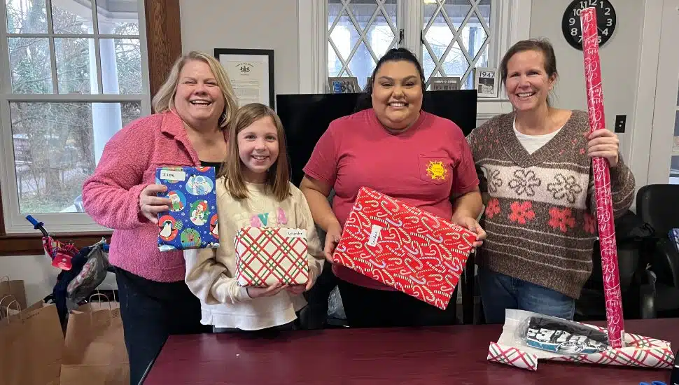 Two women and a girl hold wrapped gifts going to Chester County families. A third woman has a roll of wrapping paper about to wrap a donated gift.