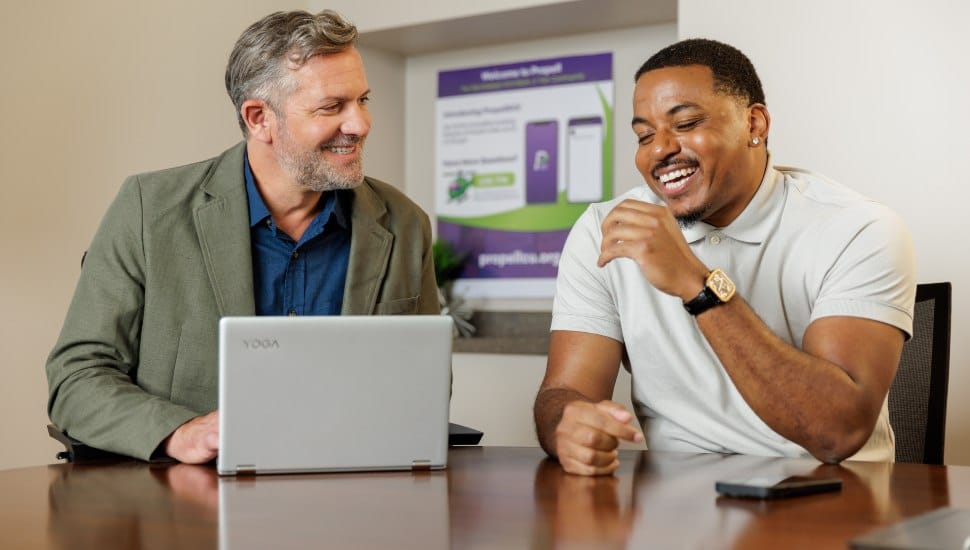 Two men sitting at a table, engaged in a friendly conversation. One man, wearing a green blazer, is using a Lenovo Yoga laptop, while the other, in a light-colored polo shirt, laughs with his hand raised. A sign in the background promotes Propell Credit Union's digital banking services.