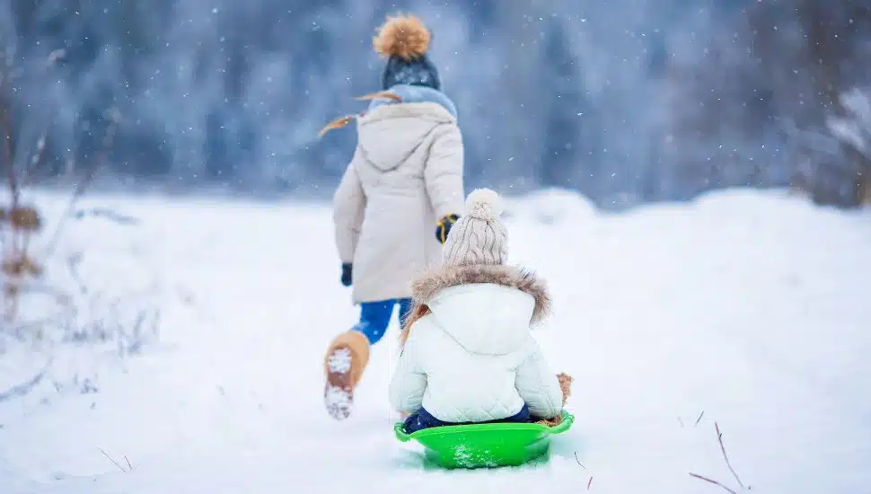 Young girl pulling another on a green sled in the snow.