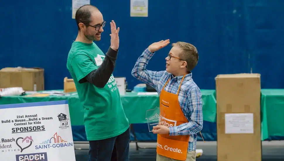 An adult in a green Habitat for Humanity t-shirt and a young boy in an orange apron high-five at the 21st Annual Build a House… Build a Dream contest. The boy holds a clear award, celebrating his achievement, while a sponsor sign and event tables are visible in the background.