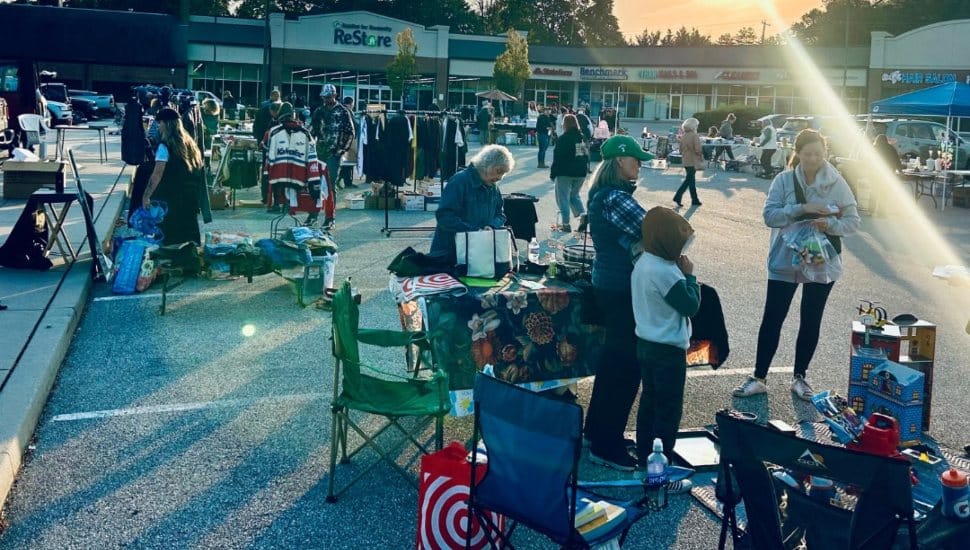 People shopping and selling various items at an outdoor yard sale in the Phoenixville ReStore parking lot, with tables, clothing racks, and merchandise set up under the morning sun.