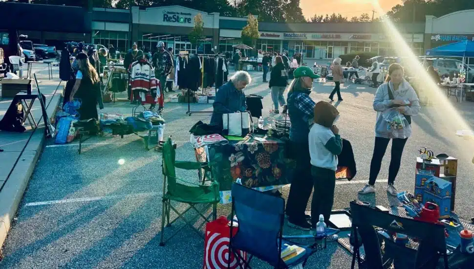 People shopping and selling various items at an outdoor yard sale in the Phoenixville ReStore parking lot, with tables, clothing racks, and merchandise set up under the morning sun.