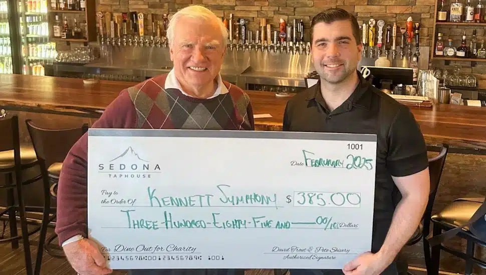 Two men stand inside Sedona Taphouse, smiling while holding an oversized check for $385 made out to Kennett Symphony. The check, dated February 2025, is part of the 'Dine Out for Charity' fundraiser. The background features a bar with various beer taps and a warm, rustic interior.