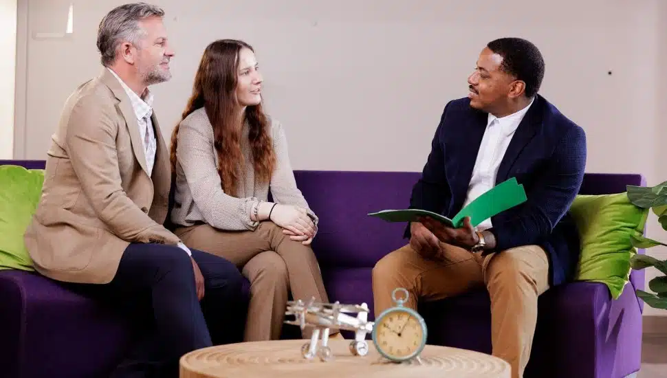 A professional financial advisor in a navy blazer and khaki pants sits on a purple couch, holding a green folder, while discussing loan options with a smiling couple dressed in business casual attire. The setting is a modern, well-lit office with green accent pillows and a decorative coffee table.