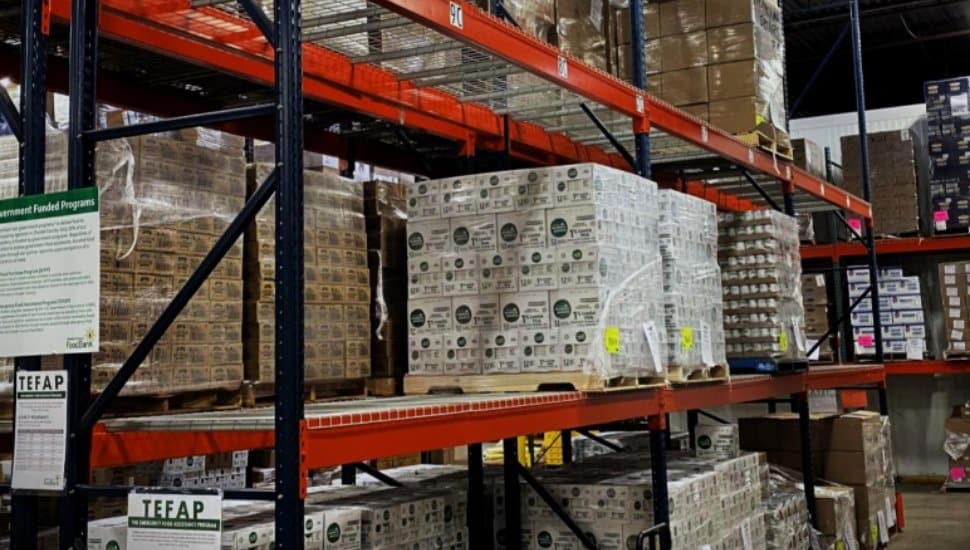 A warehouse with tall metal shelves stocked with boxes of food supplies, including TEFAP-labeled products. A green sign on the left provides information about government-funded food programs.