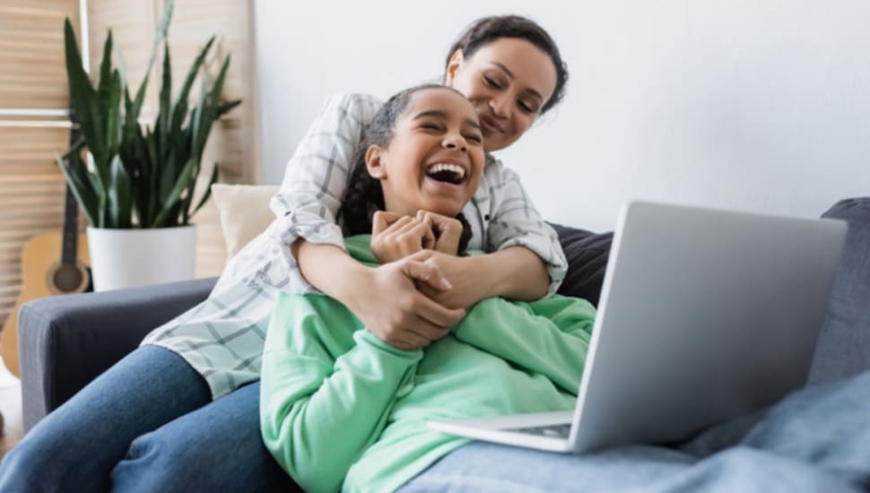 A mom and her teenage daughter share a moment while the girl is on her laptop.