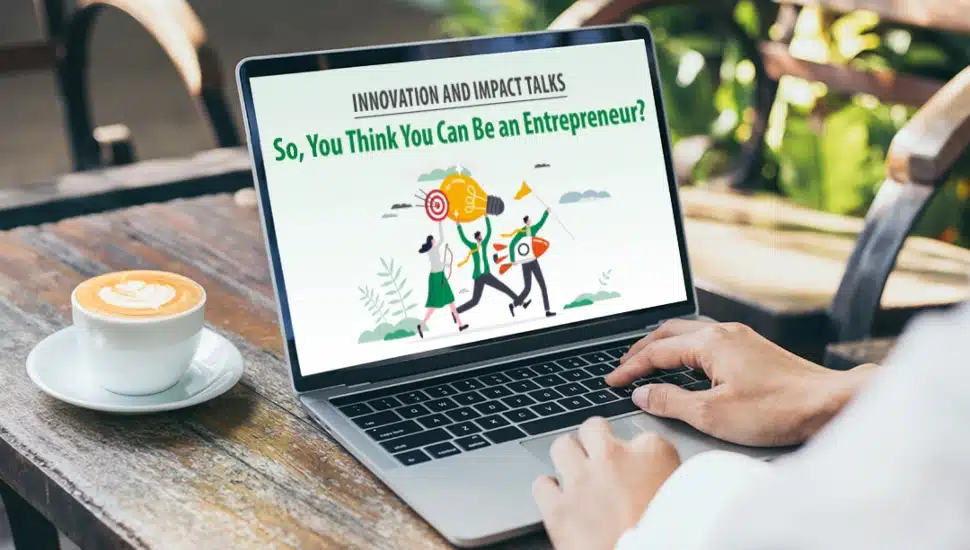 A person using a laptop at an outdoor café with a cup of coffee beside them. The laptop screen displays a promotional graphic for Wilmington University’s Innovation and Impact Talks event titled "So, You Think You Can Be an Entrepreneur?" featuring an illustration of people carrying a giant lightbulb, symbolizing innovation and entrepreneurship.