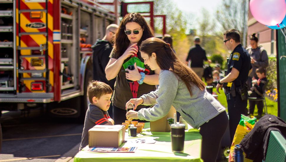 Children and families enjoy a hands-on activity at a YMCA Healthy Kids Day event. A woman helps a young boy at a craft table set up outdoors, with a fire truck and police officers visible in the background. The sunny day and colorful balloons create a festive, community-focused atmosphere.