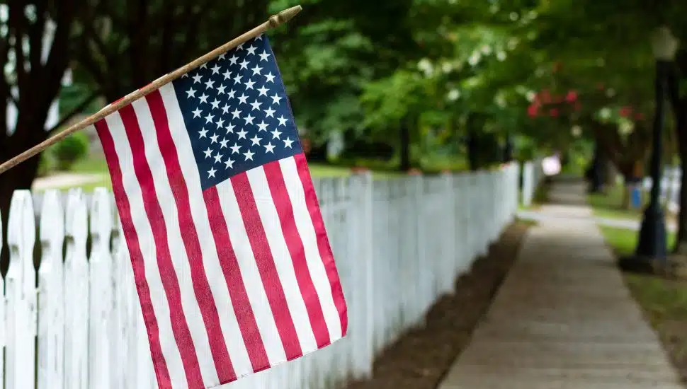 Small American flag hangs from a picket fence along the sidewalk in a rural town.