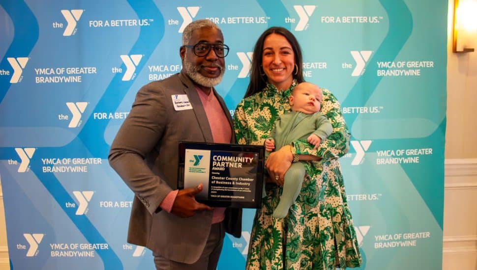 Bertram L. Lawson II, President and CEO of YGBW holding a YMCA Community Partner Award plaque. He stands beside Laura Manion, who is in a green floral dress and holding a baby. They are posing in front of a YMCA of Greater Brandywine step-and-repeat backdrop.