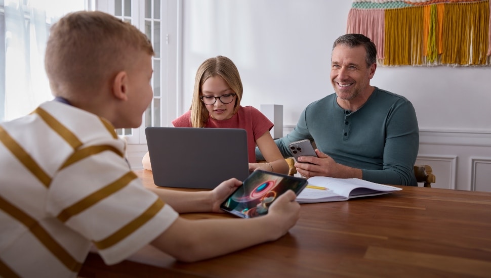 A father sits a table with daughter and son on electronics.