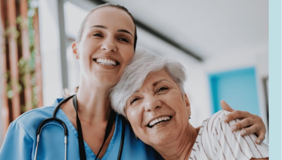 Young nurse side-hugging older woman, both are smiling.
