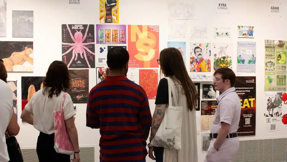 A group of people viewing student artwork displayed on a gallery wall, featuring colorful posters, paintings, and graphic designs at the AFA Graduation Exhibition at Delaware County Community College.