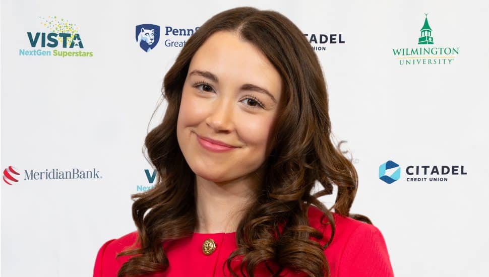 Headshot of Gina Lizzo. She has long wazy brown hair, and is smiling with her mouth closed. She wears a red blazer and her head is titled to the side.