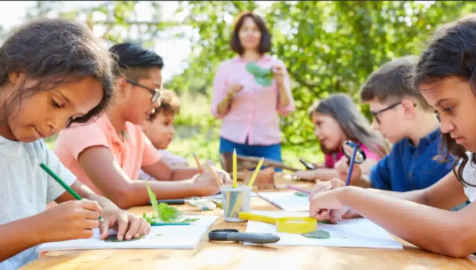 Children working on a craft project at a summer camp table outside while a counselor looks on.