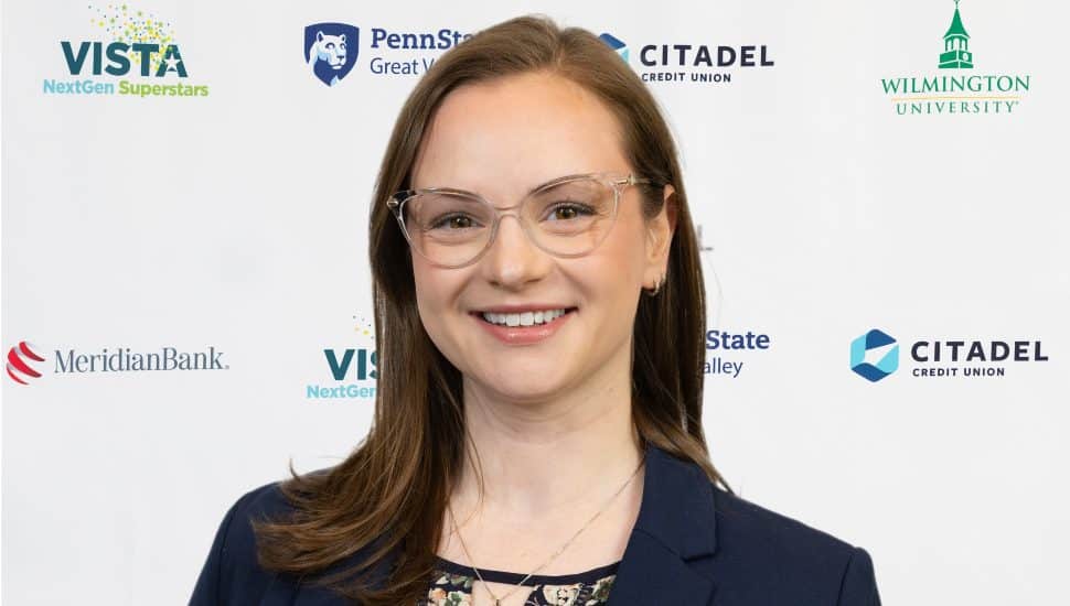 Headshot of Kate Hutelmyer posed in front of the 2025 NextGen Superstar banner. She wears clear glasses, is smiling, and has shoulder-length brown hair.