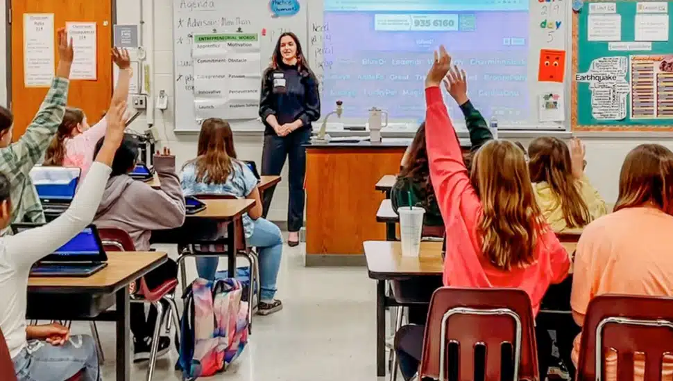 Teacher stands in front of classroom with students raising their hands.