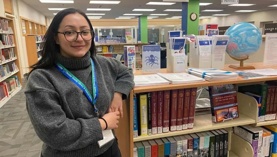 Business and Career Librarian, Mikaila Strano. She has long dark hair, wears glasses, has her arm propped on a bookshelf, and is smiling.