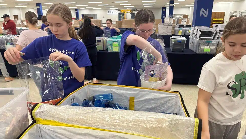 Young volunteers assemble Easter baskets at a community event, carefully placing items into clear bags. Tables filled with supplies and other participants are visible in the background, highlighting the teamwork and organization of the event.