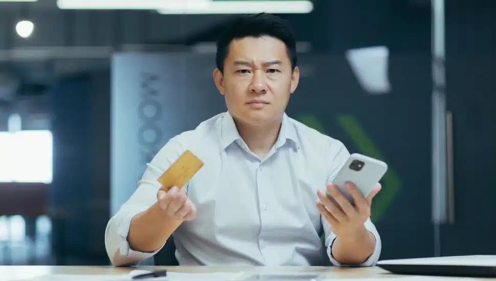 A frustrated man holding credit card and phone at a table, disgruntled with his bank.