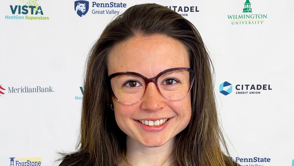 Headshot of Lydia Kirby in front of the VNS2025 banner. Lydia has shoulder-length brown hair and large classes and is smiling.