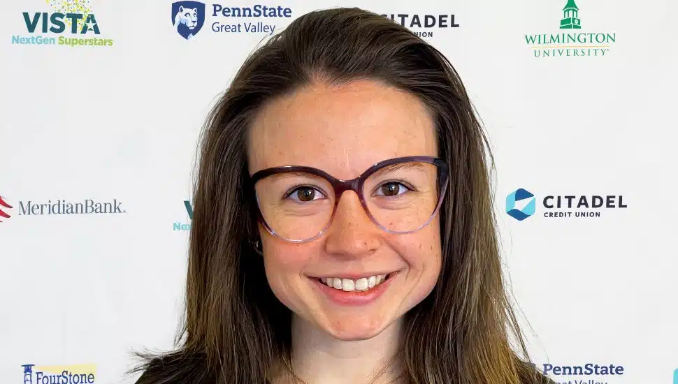 Headshot of Lydia Kirby in front of the VNS2025 banner. Lydia has shoulder-length brown hair and large classes and is smiling.
